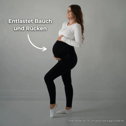 Das Bild zeigt eine schwangere Frau, die eine schwarze Umstandsmode-Leggings trägt. Sie steht in einer seitlichen Pose und hält ihren Babybauch, während ein Text „Entlastet Bauch und Rücken“ auf das Bild hinweist. Die Frau trägt ein weißes Oberteil und lächelt, während sie in einem neutralen, hellen Raum steht.