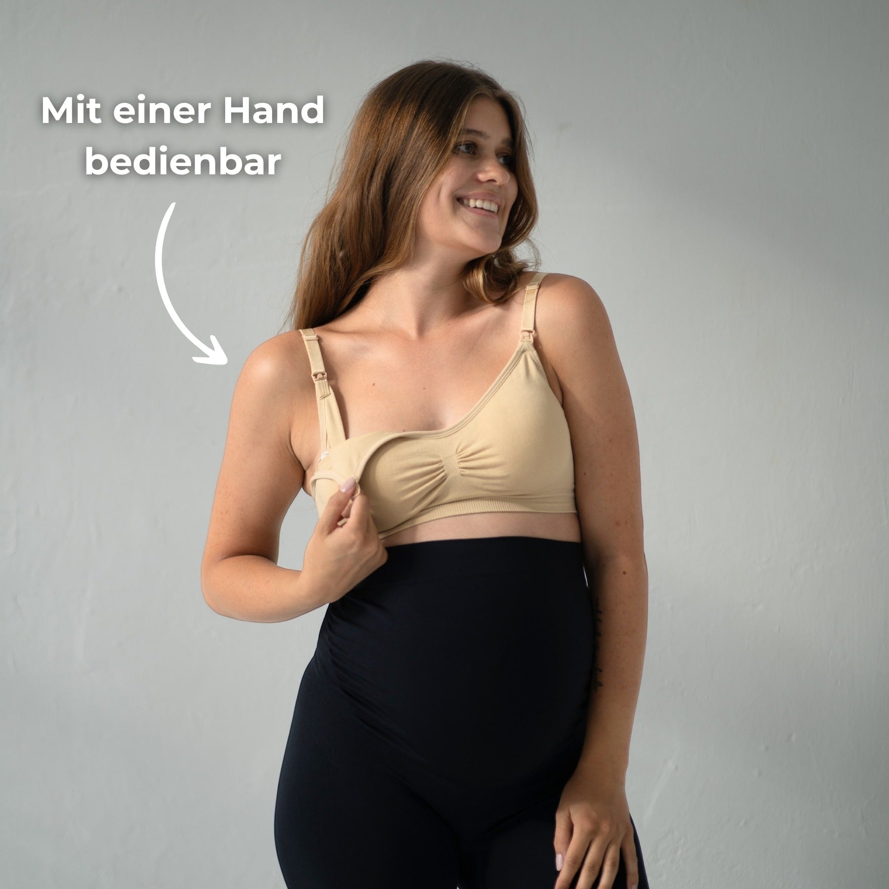 Das Bild zeigt eine werdende Mutter, die einen Still-BH trägt. Sie hebt den BH mit einer Hand, um zu zeigen, dass der Verschluss des BHs einfach mit einer Hand bedient werden kann. Der Text „Mit einer Hand bedienbar“ ist neben ihr platziert, um diesen praktischen Vorteil des BHs zu betonen. Der Hintergrund ist neutral, und die Mutter lächelt, während sie den BH anhebt.