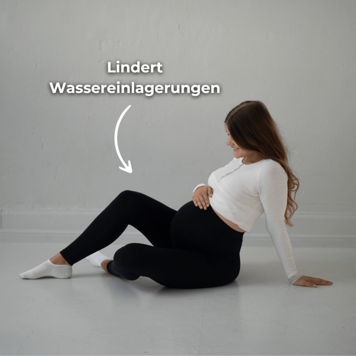 Das Bild zeigt eine schwangere Frau, die schwarze Umstandsmode-Leggings und ein weißes, langärmliges Oberteil mit einem kleinen Knopfverschluss trägt. Sie sitzt auf dem Boden, lehnt sich leicht zurück und hält ihren Babybauch mit einer Hand, während die andere Hand auf dem Boden abstützt. Der Text „Lindert Wassereinlagerungen“ zeigt, dass die Leggings dazu beiträgt, Beschwerden durch Wassereinlagerungen zu lindern. Ihre braunen Haare sind locker und fallen über ihre Schultern. Der Raum ist schlicht und hell