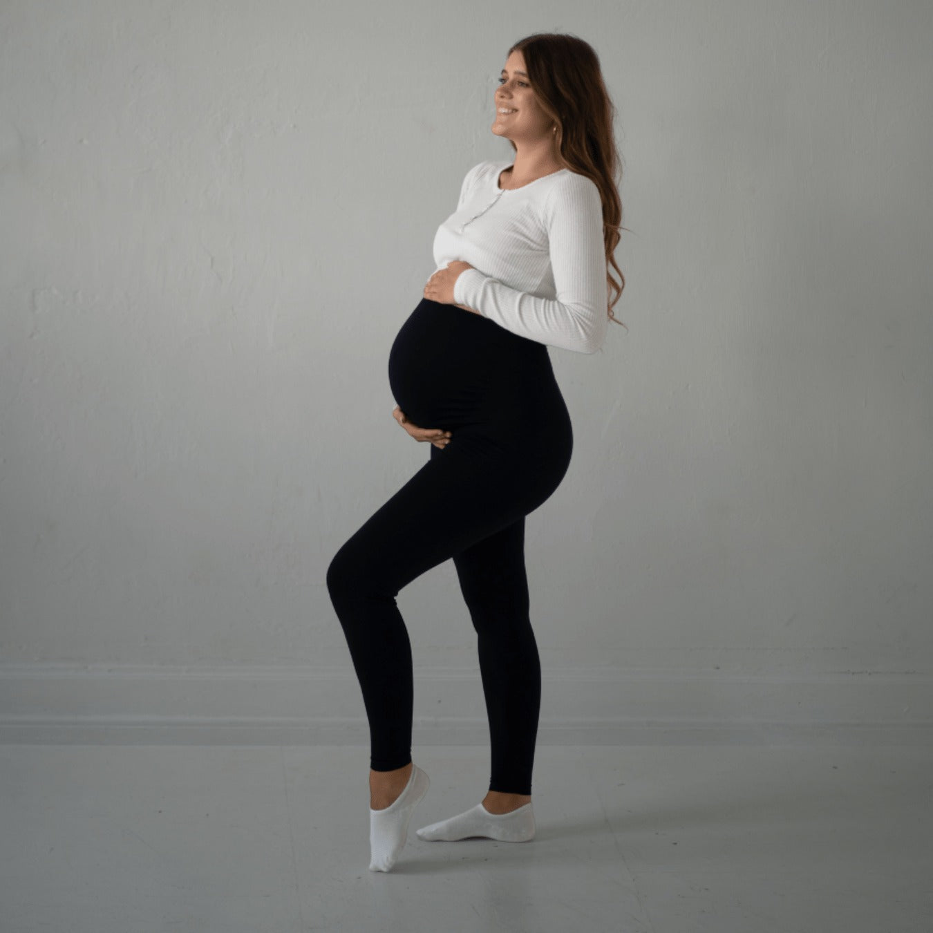 Das Bild zeigt eine schwangere Frau, die schwarze Umstandsmode-Leggings trägt, zusammen mit einem weißen, langärmligen Oberteil. Sie steht seitlich, hält ihren Babybauch mit einer Hand und lächelt. Ihre braunen Haare fallen locker über ihre Schultern. Der Hintergrund ist schlicht und hell, was die Frau und die Leggings in den Mittelpunkt stellt.