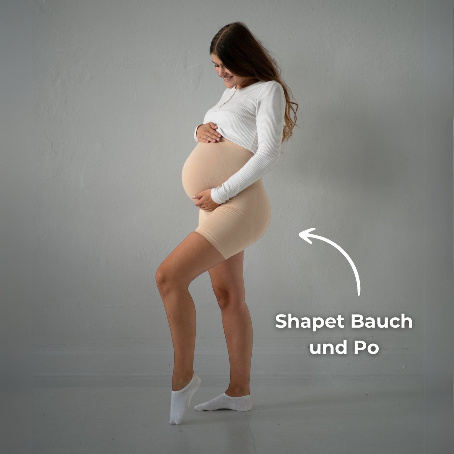 Das Bild zeigt eine schwangere Frau, die beige Umstandsmode-Shorts trägt. Sie steht in einer seitlichen Pose und hält ihren Babybauch mit einer Hand. Der Text „Shapet Bauch und Po“ weist darauf hin, dass die Shorts den Bauch und Po formen und stützen. Die Frau trägt ein weißes, langärmliges Oberteil und trägt weiße Socken. Der Hintergrund ist neutral und hell. Sie lächelt und scheint sich in der Leggings wohl zu fühlen.