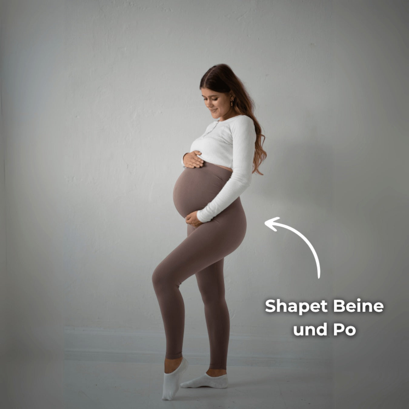 Das Bild zeigt eine schwangere Frau, die braune Umstandsmode-Leggings trägt. Sie steht in einer seitlichen Pose und hält ihren Babybauch, während ein Text „Shapet Beine und Po“ auf das Bild hinweist. Die Frau trägt ein weißes Oberteil und steht in einem neutralen, hellen Raum.