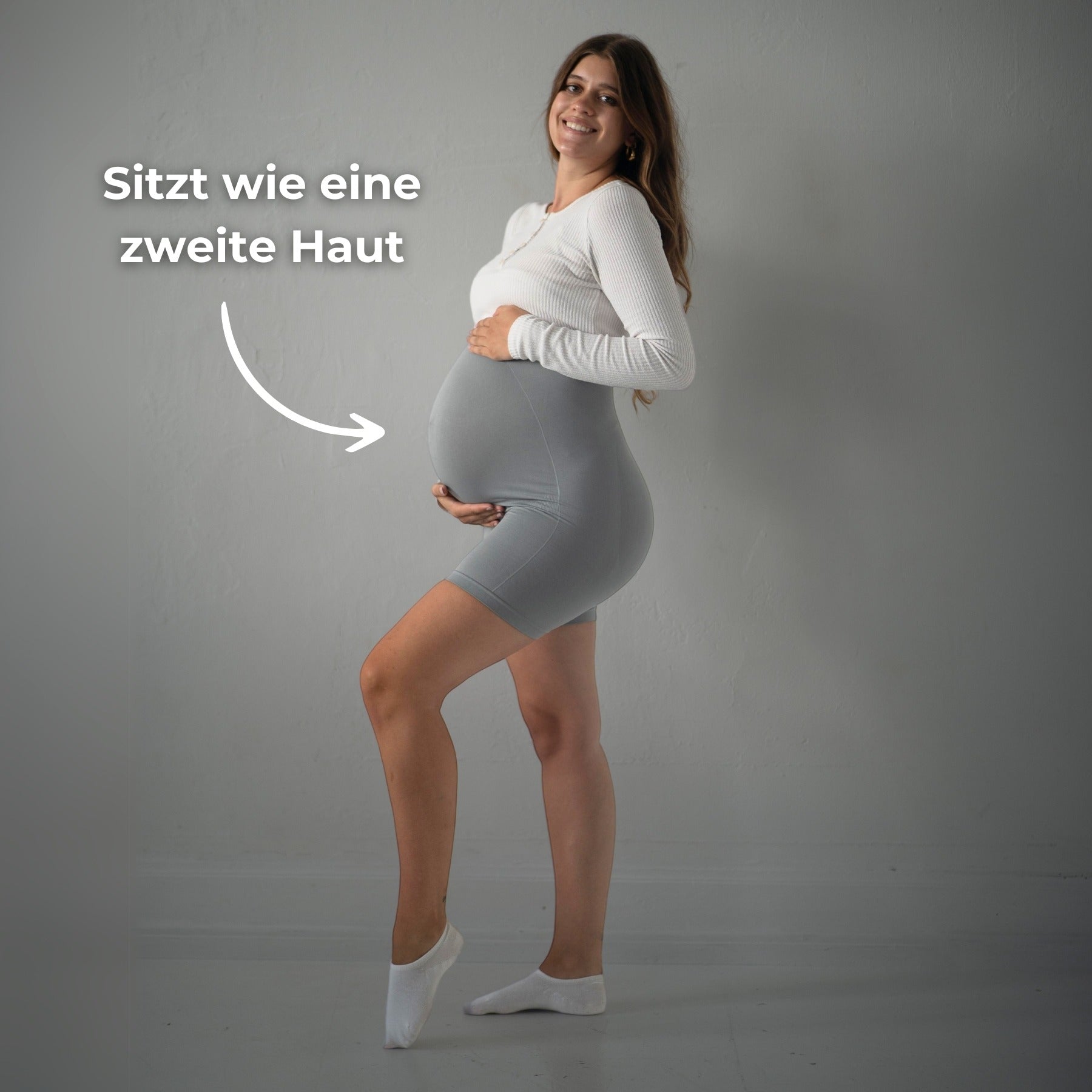 Das Bild zeigt eine schwangere Frau, die graue Umstandsmode-Shorts trägt. Sie steht seitlich und hält ihren Babybauch mit einer Hand. Der Text „Sitzt wie eine zweite Haut“ deutet darauf hin, dass die Shorts sehr gut anliegt und sich angenehm an den Körper schmiegt. Sie trägt ein weißes, langärmliges Oberteil und weiße Socken. Der Hintergrund ist neutral und die Frau lächelt, während sie in die Kamera blickt.