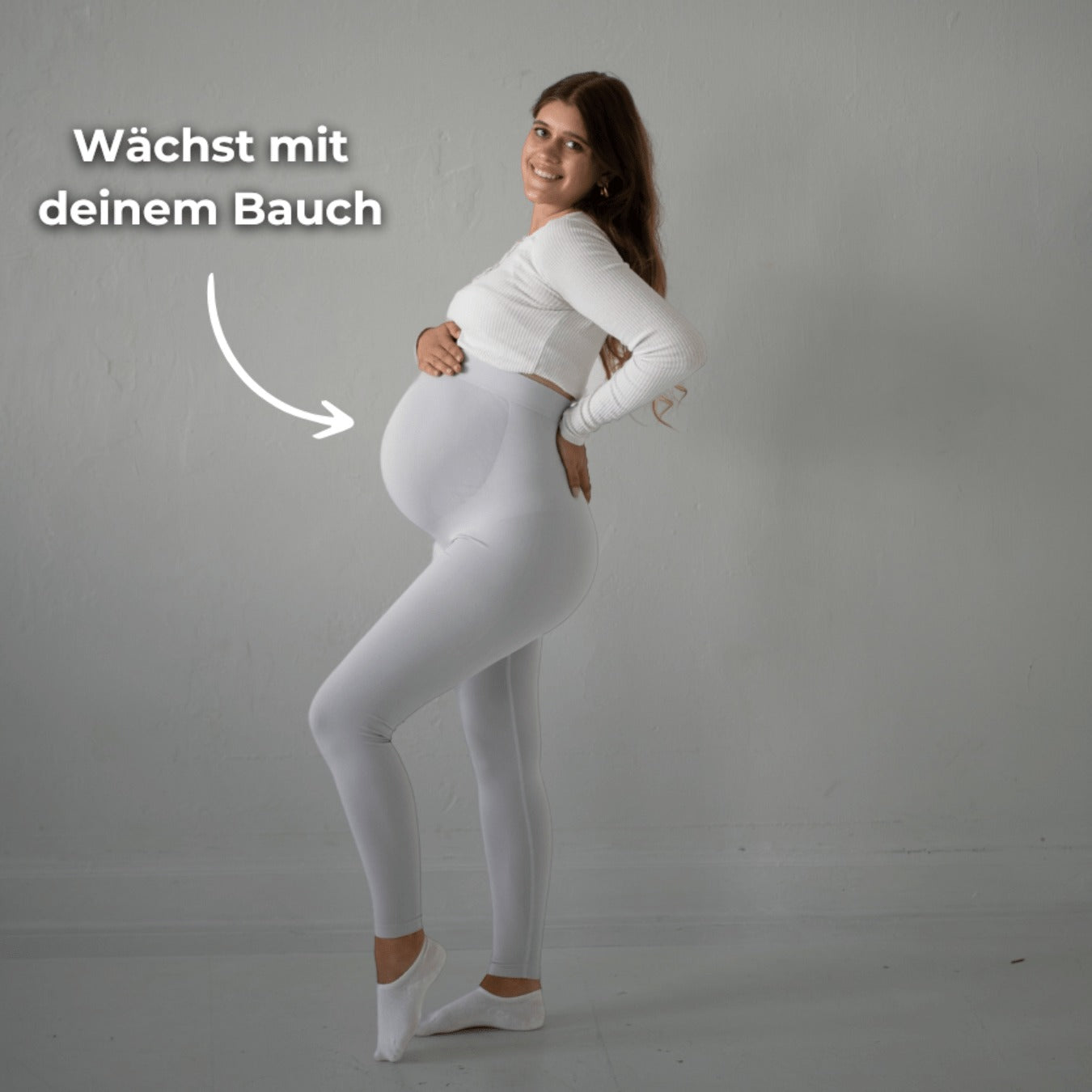 Das Bild zeigt eine schwangere Frau, die weiße Umstandsmode-Leggings trägt. Sie steht in einer seitlichen Pose, hält ihren Babybauch und lächelt. Ein Text auf dem Bild weist darauf hin, dass die Leggings „mit deinem Bauch wächst“. Sie trägt ein weißes Oberteil und befindet sich in einem neutralen, hellen Raum.