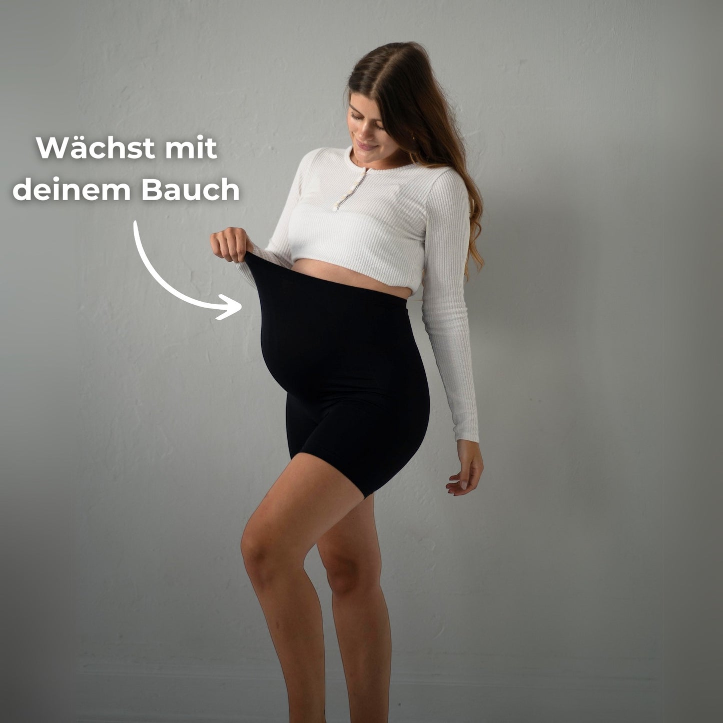 Das Bild zeigt eine schwangere Frau, die schwarze Umstandsmode-Shorts trägt. Sie steht seitlich und zieht die Shorts mit einer Hand am oberen Rand hoch, um zu zeigen, dass sie mit dem Bauch wächst. Der Text „Wächst mit deinem Bauch“ unterstreicht, dass die Shorts sich an die Veränderungen des Körpers anpasst. Die Frau trägt ein weißes, langärmliges Oberteil und hat ein freundliches Lächeln im Gesicht. Der Hintergrund ist neutral und schlicht.