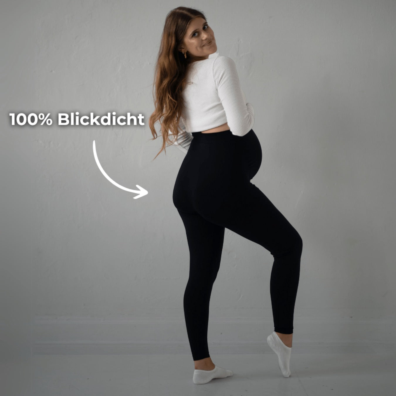 Das Bild zeigt eine schwangere Frau, die schwarze Umstandsmode-Leggings trägt. Sie steht in einer seitlichen Pose, dreht sich leicht zur Kamera und zeigt ihren Babybauch. Der Text auf dem Bild hebt hervor, dass die Leggings „100% blickdicht“ ist. Sie trägt ein weißes Oberteil und befindet sich in einem neutralen, hellen Raum.