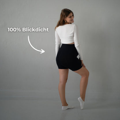 Das Bild zeigt eine schwangere Frau, die schwarze Umstandsmode-Shorts trägt. Sie steht seitlich und hebt ein Bein leicht an, während sie in die Kamera lächelt. Der Text „100% Blickdicht“ weist darauf hin, dass die Shorts vollständig blickdicht ist und eine hohe Abdeckung bietet. Die Frau trägt ein weißes, langärmliges Oberteil und weiße Socken. Der Hintergrund ist schlicht und neutral, was die Frau und die Shorts in den Fokus rückt.