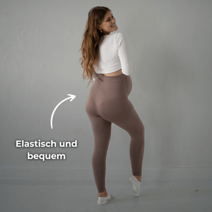 Das Bild zeigt eine schwangere Frau, die braune Umstandsmode-Leggings trägt. Sie steht seitlich und dreht sich leicht zur Kamera. Der Text „Elastisch und bequem“ auf dem Bild weist darauf hin, dass die Leggings besonders dehnbar und angenehm zu tragen ist. Sie trägt ein weißes Oberteil und steht vor einer neutralen Wand.