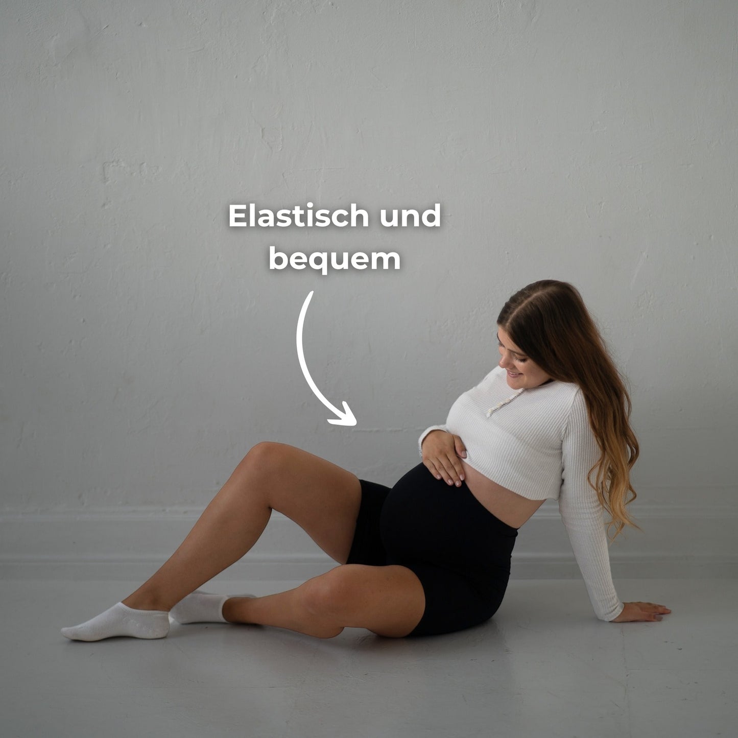 Das Bild zeigt eine schwangere Frau, die schwarze Umstandsmode-Shorts trägt. Sie sitzt auf dem Boden und hat ein Bein ausgestreckt, während sie ihren Babybauch mit einer Hand stützt. Der Text „Elastisch und bequem“ betont, wie flexibel und angenehm die Shorts ist. Sie trägt ein weißes, langärmliges Oberteil und weiße Socken. Der Hintergrund ist schlicht und neutral, was die Frau und die Shorts in den Mittelpunkt stellt. Die Frau hat ein Lächeln im Gesicht und wirkt entspannt.