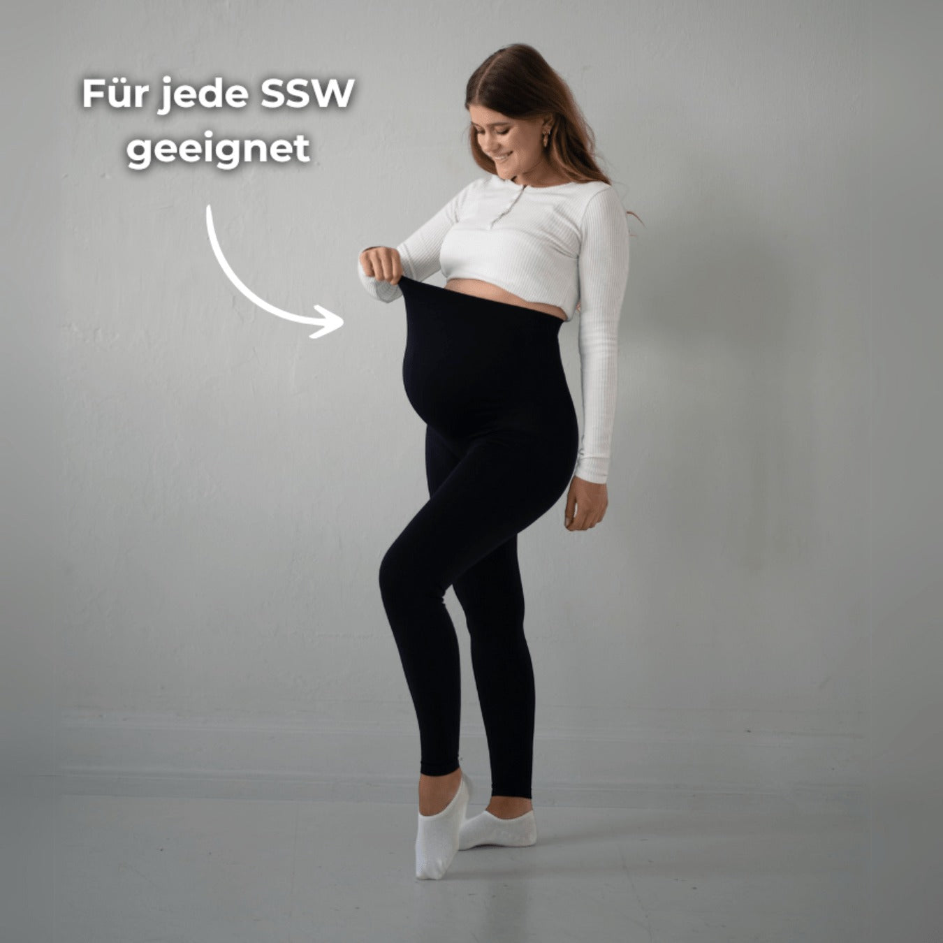 Das Bild zeigt eine schwangere Frau, die schwarze Umstandsmode-Leggings trägt. Sie hält die Leggings an ihrem Babybauch, um zu zeigen, dass sie „für jede SSW geeignet“ ist. Sie trägt ein weißes Oberteil und steht vor einer neutralen Wand.