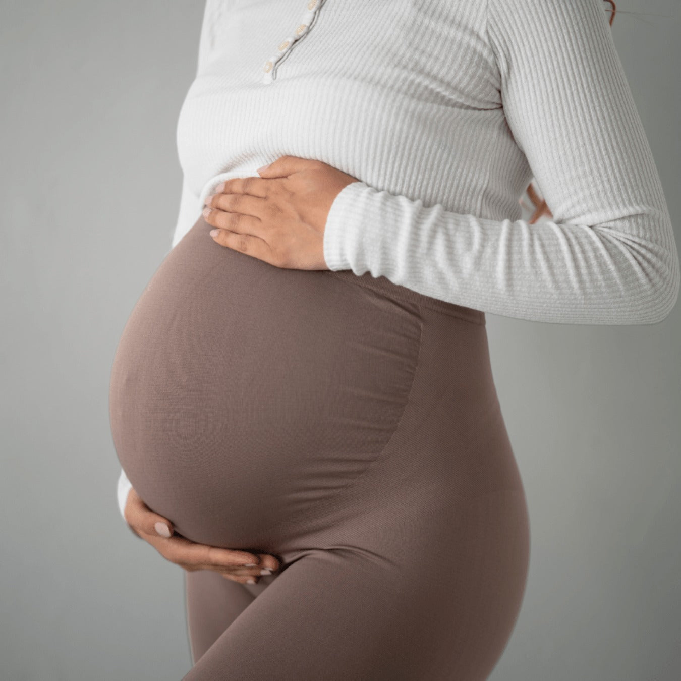 Das Bild zeigt eine schwangere Frau, die braune Umstandsmode-Leggings trägt. Sie hält ihren Babybauch mit einer Hand, während sie ein weißes Oberteil trägt. Das Bild konzentriert sich auf die Leggings und den Babybauch der Frau.