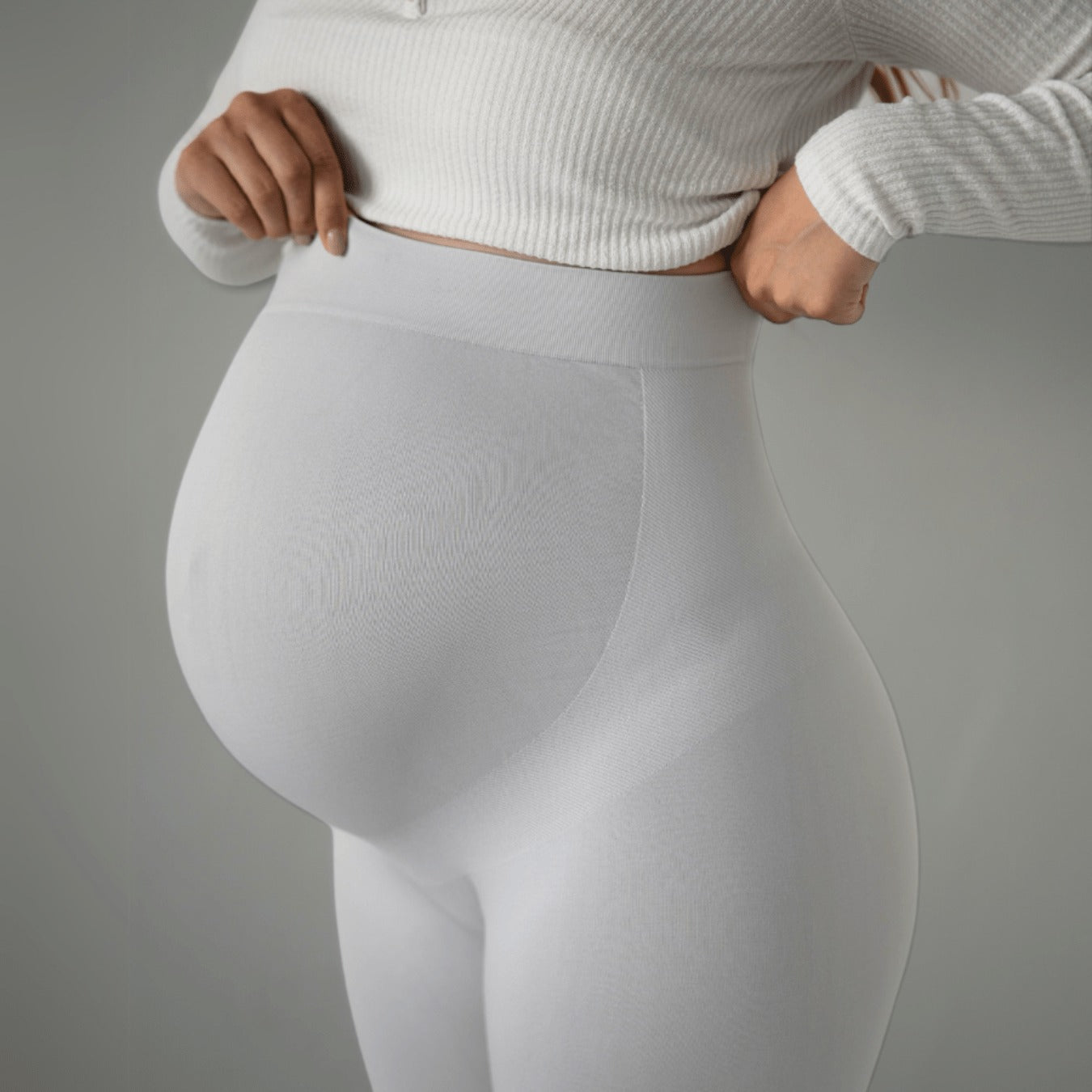 Das Bild zeigt eine schwangere Frau, die weiße Umstandsmode-Leggings trägt. Sie zieht die Leggings am oberen Rand leicht hoch, um den dehnbaren Stoff zu zeigen, der sich ihrem wachsenden Babybauch anpasst. Sie trägt ein weißes Oberteil und das Bild fokussiert auf den Bauchbereich.